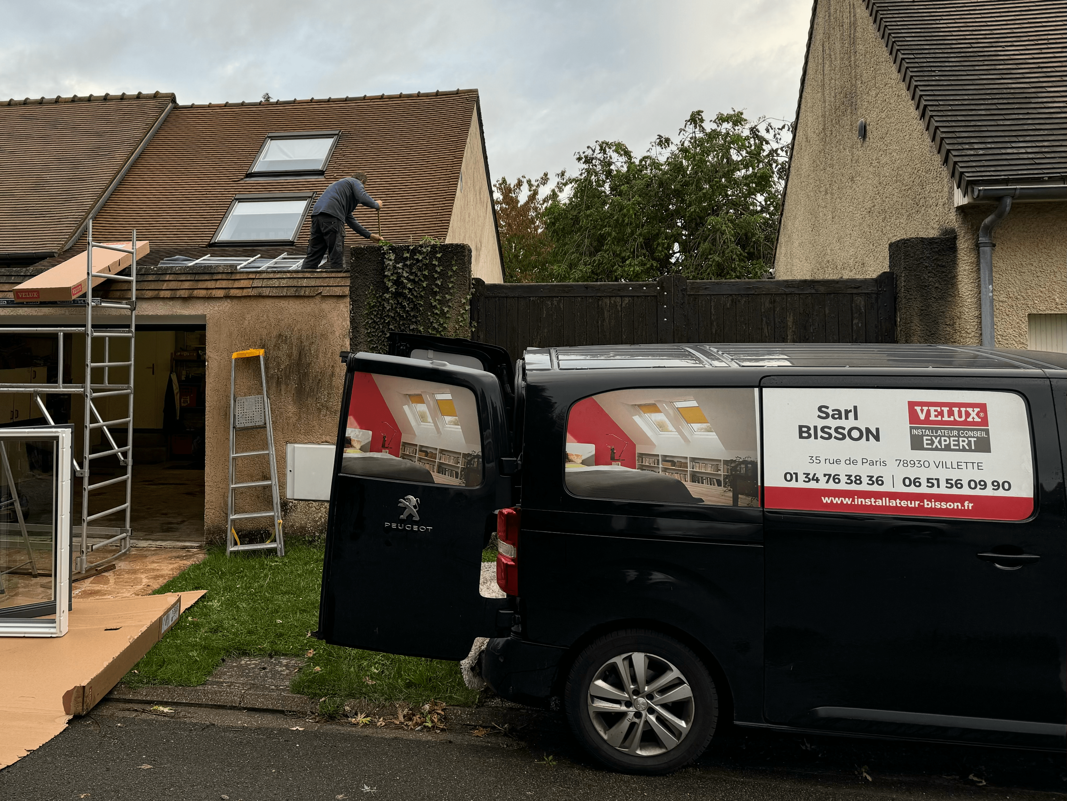 Un professionnel réalise la pose Velux en renovation sur une toiture. L'installation de plusieurs fenêtres de toit Velux est en cours pour moderniser et améliorer l'éclairage naturel de la maison. Le véhicule de l'entreprise "SARL Bisson", spécialisé en pose et renovation de fenêtres de toit Velux, est stationné au premier plan.