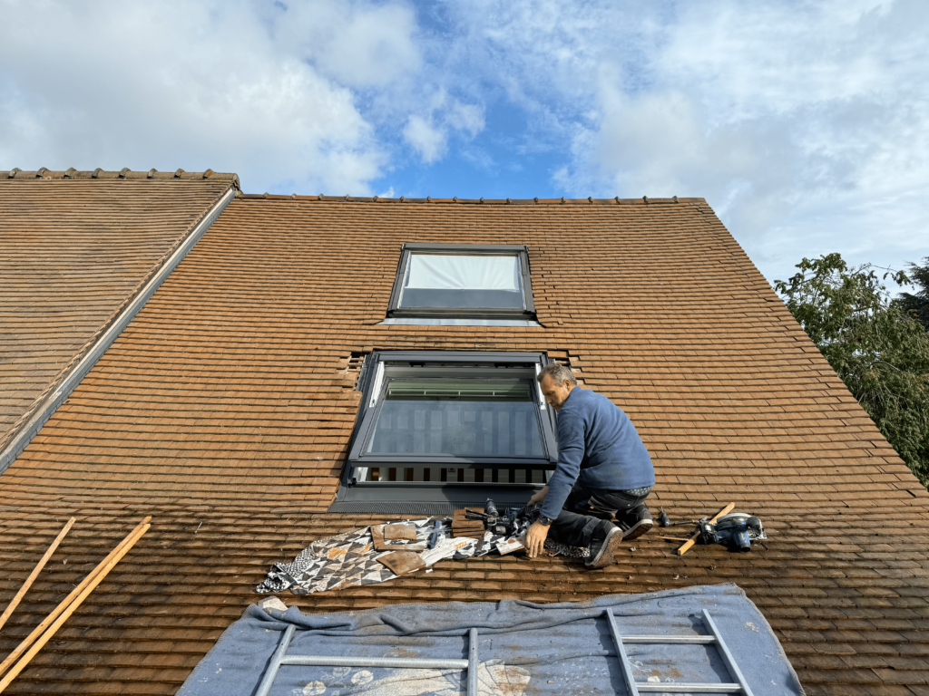 L'installation d'une fenêtre de toit Velux en renovation est réalisée par un professionnel sur une toiture en tuiles plaques, avec un encadrement soigneusement ajusté pour garantir une étanchéité parfaite et une intégration harmonieuse.